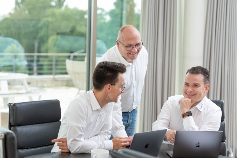 Drei Männer in einem modernen Büro, die gemeinsam an Laptops arbeiten und lächelnd miteinander sprechen.